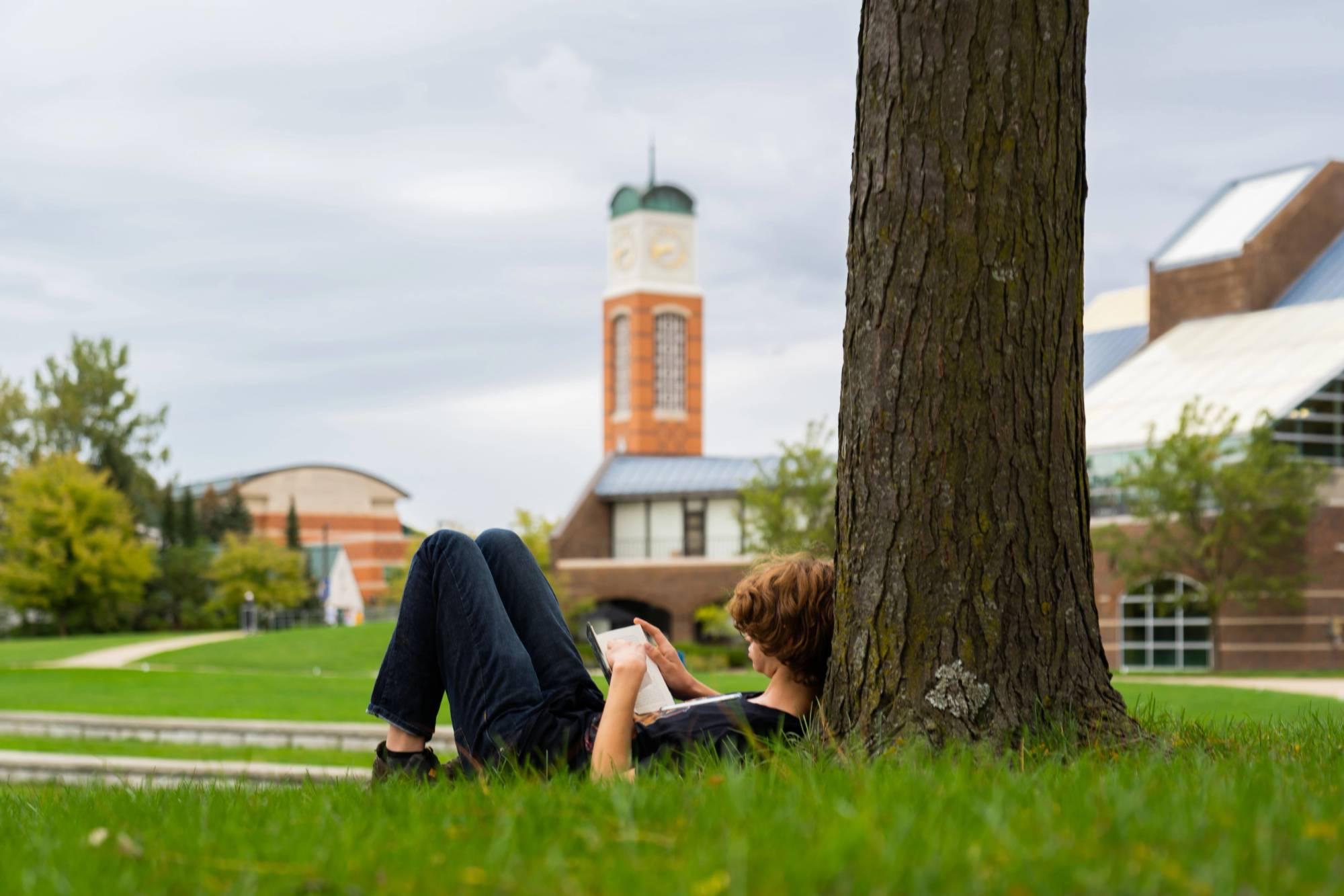 A person works on an iPad while laying under a tree near the Cook Carillon Tower